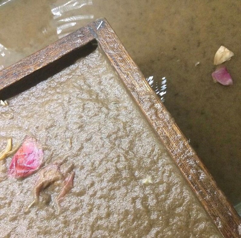Puede incluir: Primer plano de un marco de madera que rodea una hoja de papel hecho a mano. El papel es de color marr&oacute;n claro y tiene una textura rugosa. Hay peque&ntilde;os trozos de flores y hojas secas incrustados en el papel.