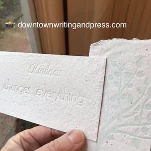 May include: A white card with the text "Kindness changes everything" printed on it. The card is made of recycled paper and has a textured surface.