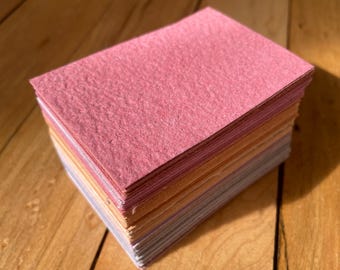 May include: A stack of colorful felt squares in shades of pink, orange, and lavender. The felt squares are stacked neatly on a wooden surface, with the top layer being a vibrant pink.