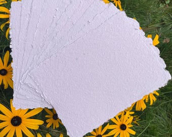 May include: A stack of rectangular sheets of light lavender handmade paper with rough, deckled edges. The paper has a visible fibrous texture and is layered in a fan shape, showcasing the uneven, organic borders of each individual sheet.