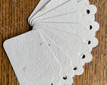 May include: A stack of off-white, scalloped-edge tags with small holes at the top. The tags have a textured surface and are arranged in a fan shape, resting on a wooden surface. The tags are blank and ready for use.