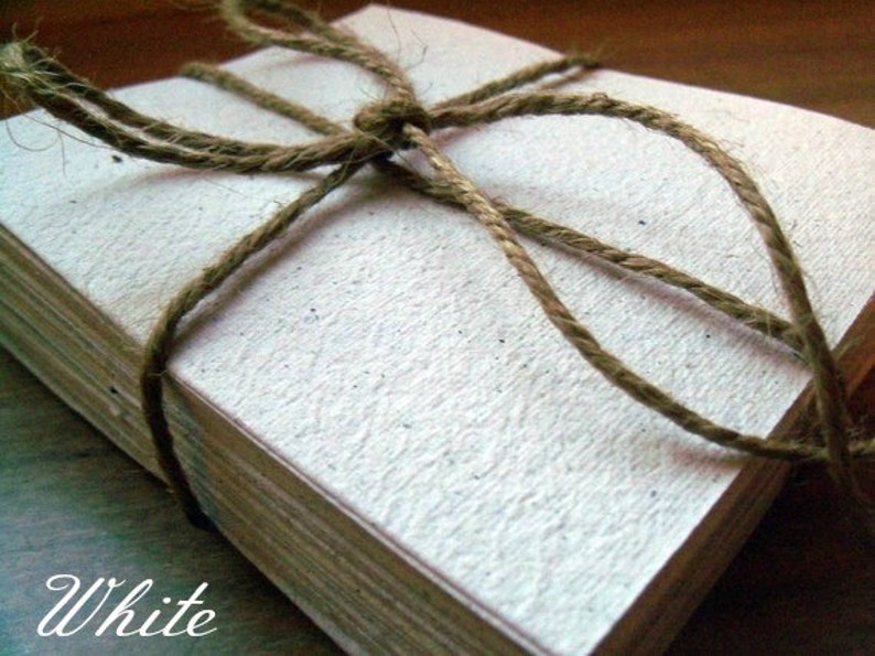 Op de afbeelding: Een stapel witte vellen papier die met touw aan elkaar zijn gebonden. Het papier heeft een textuur. Het woord "White" staat in cursief onder de stapel papier.
