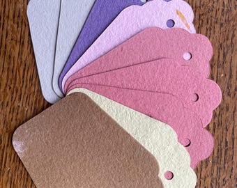 May include: A fan of scalloped-edge paper tags in various colors, including brown, pink, purple, and cream. Each tag has a small hole at the top for stringing. The tags are arranged on a wooden surface.