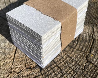 May include: A stack of small, rectangular white paper cards bound with a brown paper band. The cards have a textured surface and are neatly stacked. The image is taken in natural light.