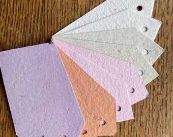 May include: A collection of colorful paper tags fanned out on a wooden surface. The tags are in shades of lavender, peach, pink, and beige, each with a small hole at the top. The tags are likely for crafts or gift giving.