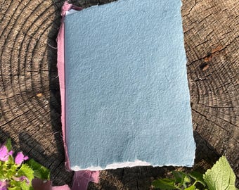May include: A rectangular notebook with a textured, light blue cover featuring rough, deckled edges. The spine is bound with a dusty rose-colored fabric ribbon that extends beyond the top and bottom edges of the book cover.