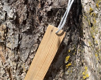 May include: A rectangular wooden bookmark with a light brown finish and visible wood grain. A light gray suede cord is attached to the top of the bookmark. The bookmark is displayed against a textured tree trunk.