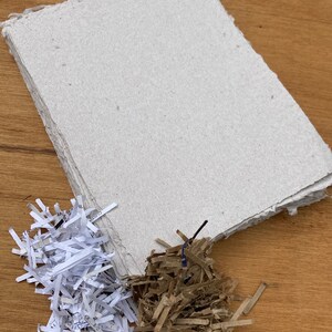 Pu&ograve; includere: Un mucchio di fogli di carta fatti a mano da carta riciclata, con carta bianca tritata e cartone marrone tritato su una superficie di legno.