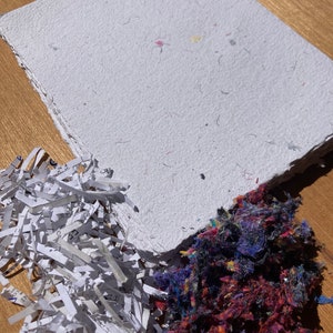 May include: A white sheet of handmade paper with flecks of color. The paper is surrounded by piles of shredded paper and fabric scraps.