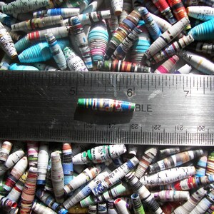 May include: A close-up of a ruler measuring a colorful, rolled paper bead. The bead is approximately 1 inch long and is made from recycled paper. The bead is surrounded by a variety of other colorful, rolled paper beads.