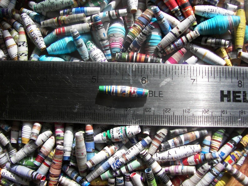 May include: A close-up of a ruler measuring a colorful, rolled paper bead. The bead is approximately 1 inch long and is made from recycled paper. The bead is surrounded by a variety of other colorful, rolled paper beads.