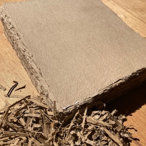 May include: A close-up of a brown cardboard sheet with a rough, textured surface. The sheet is surrounded by shredded brown paper.