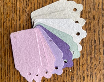 May include: A collection of pastel-colored paper tags with scalloped edges and small holes for stringing. The tags are in shades of lavender, purple, green, and off-white, arranged on a wooden surface. Ideal for crafting or gift-giving.