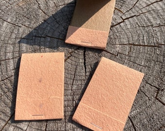 May include: Three small, brown paper notebooks with a stitched binding. The notebooks are closed and lying on a wooden surface.