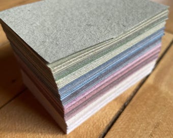May include: A stack of rectangular paper cards in various pastel colors. The cards are stacked neatly on a wooden surface. The top card is a light gray color. The edges of the cards show a spectrum of colors, including pink, blue, and green.