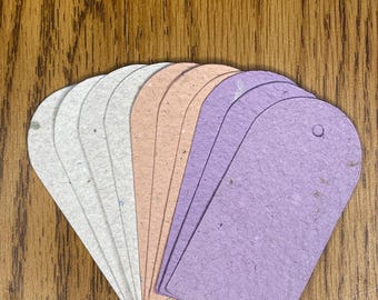 May include: A collection of blank, teardrop-shaped tags in various pastel colors, including white, light gray, peach, and lavender. Each tag has a small hole at the top. The tags are arranged on a wooden surface.