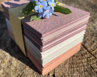 May include: A stack of square, multi-colored paper cards bound with a yellow band. A small bouquet of blue forget-me-nots with yellow centers and green leaves rests on top of the cards. The cards are arranged on a weathered wooden surface.