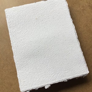 May include: A white handmade paper sheet with a rough, textured surface. The paper has a slightly irregular shape and edges.