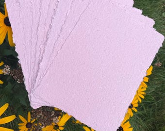 May include: A stack of rectangular sheets of light pink handmade paper with rough, deckled edges. The paper has a visible fibrous texture throughout the surface, and the sheets are layered slightly offset to show the thickness of the stack.