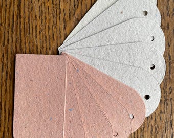 May include: A fan of teardrop-shaped paper tags in two colors: light pink and off-white. Each tag has a small hole at the top. The tags are arranged on a wooden surface, showcasing their texture and design.