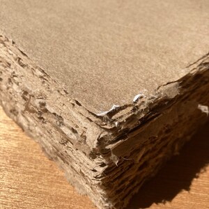 Może przedstawiać: Stos brązowych arkuszy papieru z chropowatymi krawędziami, tworzący teksturalną powierzchnię. Papier jest warstwowy i wydaje się być ręcznie robiony.