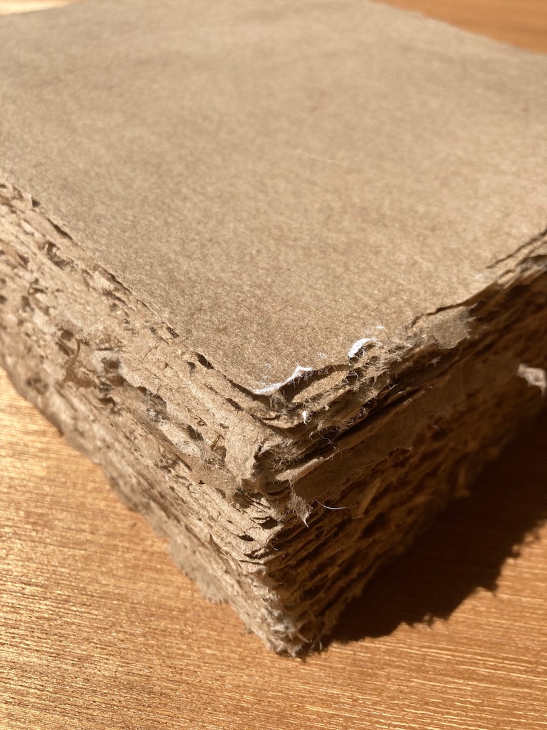 Może przedstawiać: Stos brązowych arkuszy papieru z chropowatymi krawędziami, tworzący teksturalną powierzchnię. Papier jest warstwowy i wydaje się być ręcznie robiony.