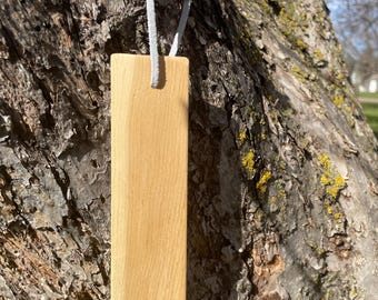 May include: A rectangular wooden bookmark with a light brown finish hangs from a gray suede cord. The bookmark is suspended against a textured tree trunk background, with a glimpse of a sunny day.