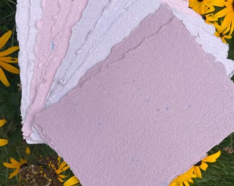 May include: A stack of rectangular handmade paper sheets with rough, deckled edges. The paper features a gradient of soft pastel colors, ranging from light pink and pale lavender to a deeper mauve, with small blue flecks embedded in the texture.