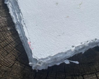 May include: A stack of thick, white, handmade paper sheets with rough, deckled edges. The surface of the top sheet is textured and contains small, visible natural fibers and a faint red speck, showing a raw, fibrous, and unrefined paper quality.