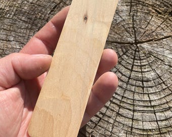 May include: A rectangular, light-colored wooden bookmark with rounded corners. The bookmark is held in a hand, showcasing its smooth, natural wood grain. The wood has a small knot. The background is a weathered tree stump.