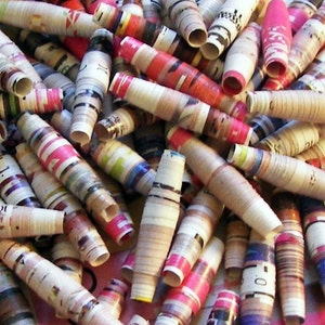 May include: A pile of colorful paper beads made from rolled up magazine pages. The beads are various lengths and have a variety of patterns and colors, including red, blue, green, and yellow.