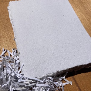 Pu&ograve; includere: Un foglio di carta bianca fatto a mano con una superficie ruvida e testurizzata. La carta &egrave; appoggiata su una superficie di legno con carta triturata in primo piano.