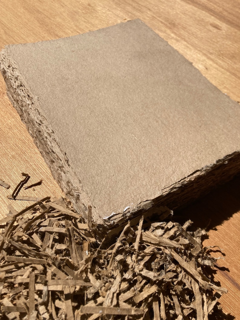 Może przedstawiać: Zbliżenie na brązowy arkusz kartonu o szorstkiej, fakturowanej powierzchni. Arkusz jest otoczony posiekanym brązowym papierem.