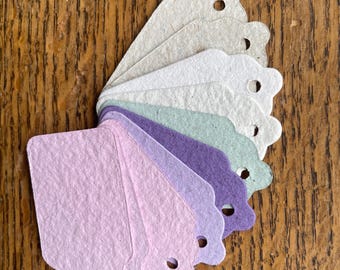 May include: A fan of paper tags in various pastel colors, including lavender, purple, and green. Each tag has a scalloped edge and a small hole at the top. The tags are arranged on a wooden surface.