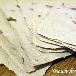 May include: A stack of handmade paper sheets with brown floral inclusions. The paper has a rough, textured surface.