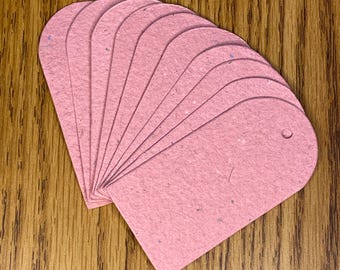 May include: A stack of pink, arched-top tags with a small hole at the top for stringing. The tags are made of a textured paper material and are arranged in a fan shape on a wooden surface.