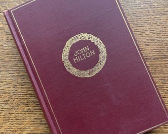 May include: A hardcover book with a burgundy cover and gold trim. The title "John Milton" is displayed in gold lettering within a circular design on the front cover. The book is resting on a wooden surface.