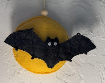 May include: A black felt bat with white eyes and beaded accents is centered over a yellow felt circle. The Halloween decoration is suspended by a natural twine string and a wooden bead. The background is a textured white wall.