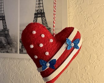 May include: Red heart-shaped ornament with white pearl accents and two blue glitter bows. The ornament has white and silver stripes and is suspended by a red and white string. The ornament is likely for Valentine's Day or a similar occasion.