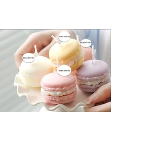 Può includere: Una collezione di quattro candele a forma di macaron nei colori pastello: giallo, rosa, bianco e lavanda, ognuna con un piccolo stoppino bianco. Le candele sono disposte su un piatto bianco e rosa con bordi smerlati. La parola "macarons" è scritta sull'immagine.