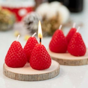 Peut inclure: Plusieurs bougies rouges en forme de fraise, dont une allumée avec une flamme jaune, sont présentées sur de petits dessous de verre en bois. Les bougies ont une surface texturée, imitant les fraises. L'arrière-plan est flou, avec d'autres objets visibles.