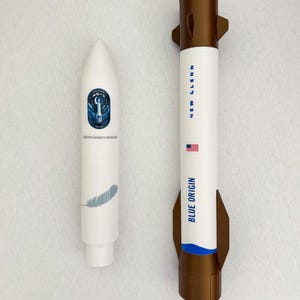 May include: Two model rockets, one white with a blue logo and a feather graphic, the other white and brown with "BLUE ORIGIN" and "NEW GLENN" printed on the side, and a small American flag.