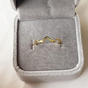 May include: A gold-colored ring with a wavy design, set in a gray velvet jewelry box. The ring's shape resembles a stylized mountain range. The ring is a delicate piece of jewelry.