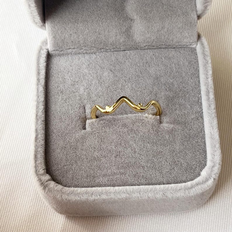 May include: A gold-colored ring with a wavy design, set in a gray velvet jewelry box. The ring's shape resembles a stylized mountain range. The ring is a delicate piece of jewelry.