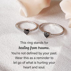 Puede incluir: Dos anillos de plata con una piedra negra en forma de corazón y pequeñas piedras transparentes. Los anillos están sobre una superficie blanca. El texto de la imagen dice: "This ring stands for healing from trauma."