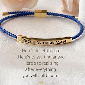 May include: A blue cord bracelet with a gold bar charm that reads "FOCK IT AND BEGIN AGAIN". The bracelet is a reminder to let go, start anew, and realize that you will still bloom after everything.