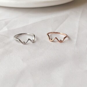 May include: Two minimalist rings. One is silver-toned, the other is rose gold. Both rings feature a wavy design with small, round accents. The rings are displayed on a white, textured surface, showcasing their simple elegance.
