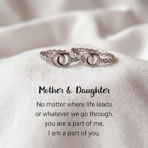 Könnte beinhalten: Zwei silberne Ringe mit ineinandergreifenden Kreisen, auf einer weißen Satin-Oberfläche. Die Ringe sind mit kleinen, funkelnden Steinen besetzt. Der Text darunter lautet "Mother & Daughter" und eine sentimentale Botschaft.