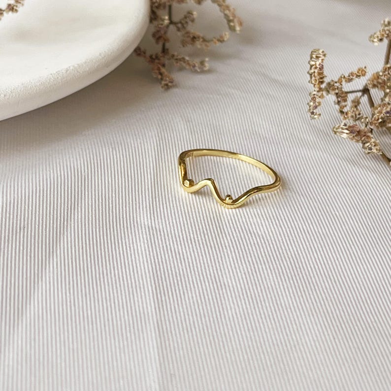 May include: A delicate gold ring with a unique, abstract design. The ring features a wavy line with two small, round accents. The ring is set against a white background with subtle textures and dried floral accents.