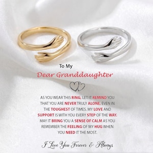 May include: Two rings, one gold-toned and one silver-toned, each featuring a design of embracing hands. The rings are displayed on a white surface with text that reads "To My Dear Granddaughter" and a loving message.